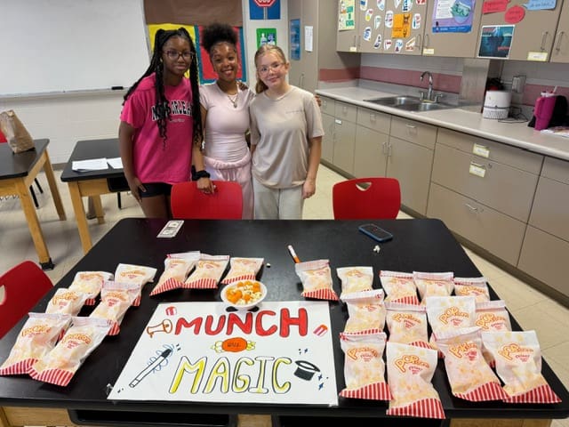 Three women standing behind a table with a sign and snack bags.