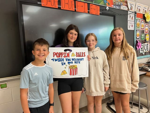 Three smiling students holding a popcorn-themed award in a classroom.
