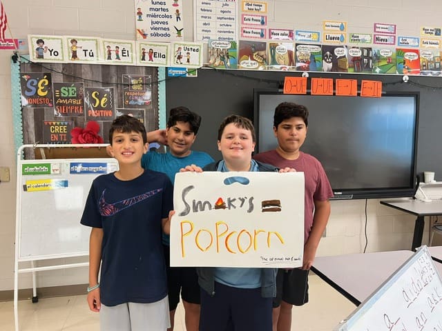 Four students holding a sign that says 'Smart Popcorn' in a classroom.