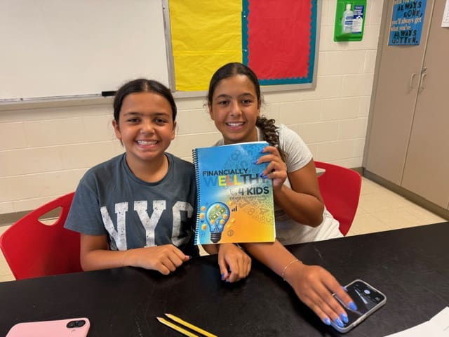 Two kids smiling and holding a colorful book in a classroom.