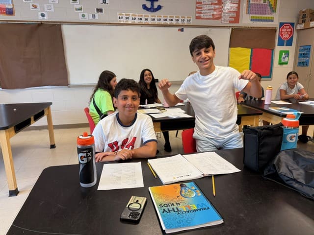 Two students smiling in a classroom with books and papers on the desk.