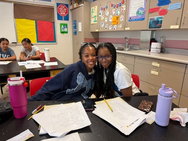 Two students smiling together in a classroom with papers on the desk.