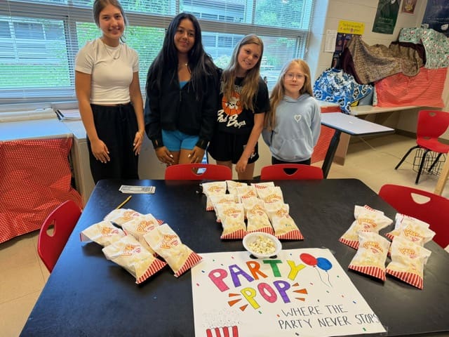 Four girls standing behind a table with popcorn bags and a colorful 'Popcorn' sign.