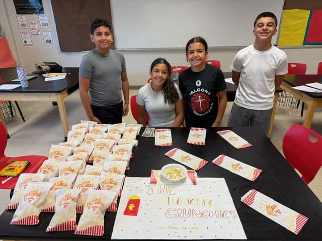 Four kids standing behind a table filled with food boxes and a birthday cake.