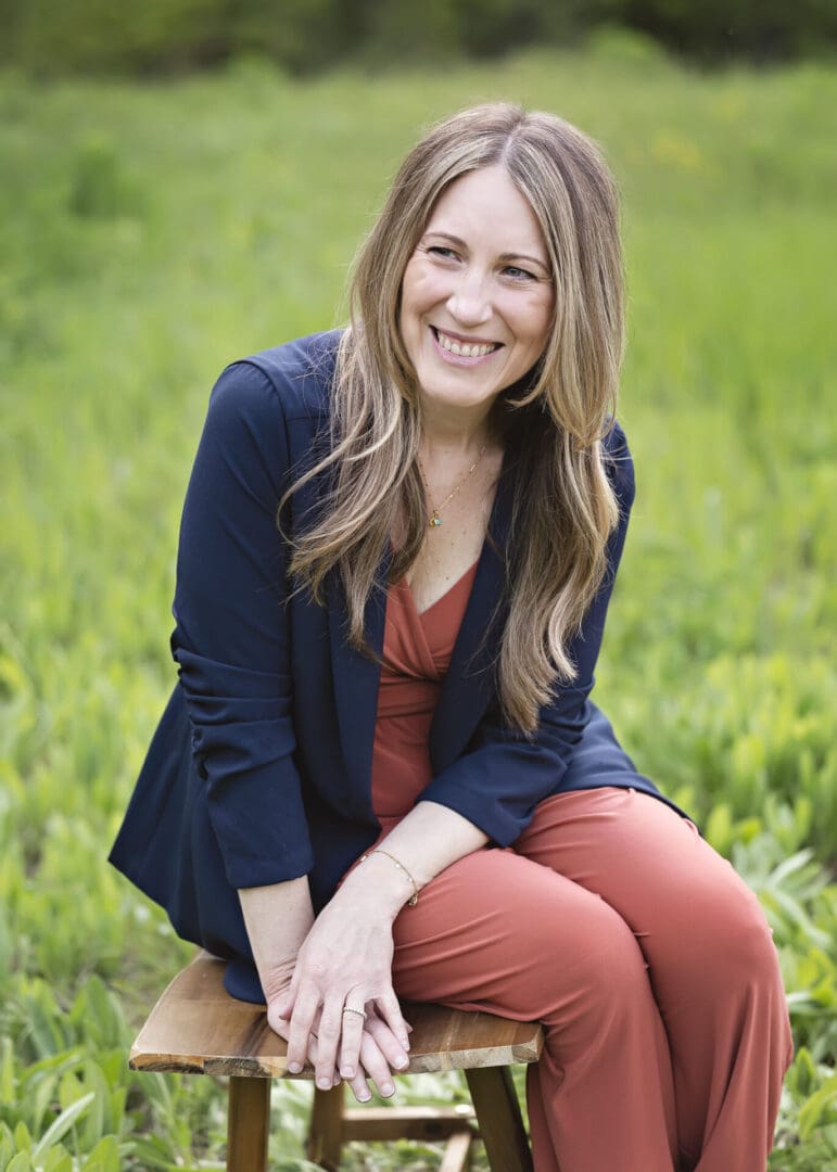A smiling woman sitting outdoors on greenery, wearing a navy blazer and rust-colored pants.