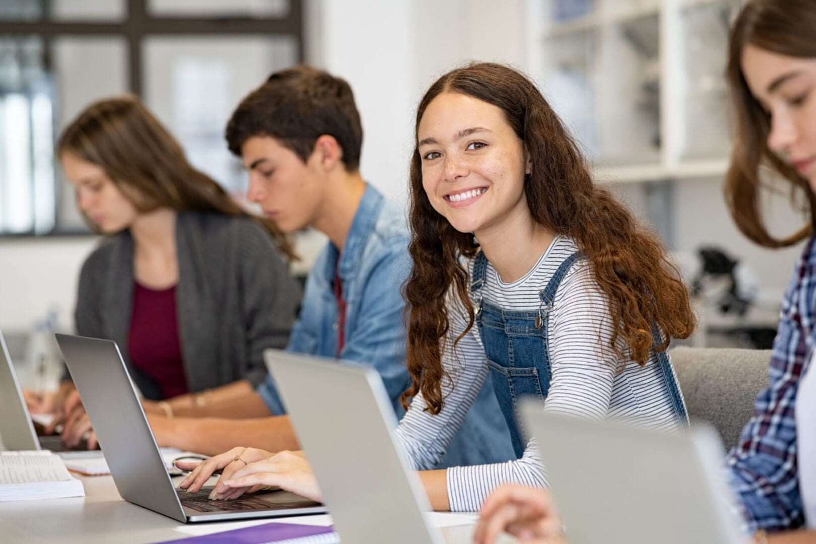 Smiling teenage girl using laptop in classroom with peers.